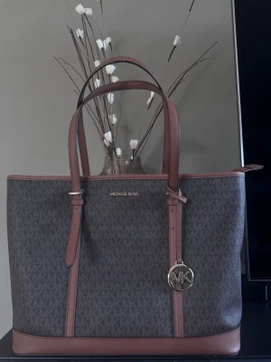 Brun Michael Kors handväska - Snygg handväska från Michael Kors i brunt med MK-logga över hela väskan. Väskan har bruna läderdetaljer, justerbara handtag och ett guldfärgat MK-hänge. Perfekt storlek för att få plats med allt du behöver. Klassisk och stilren design.