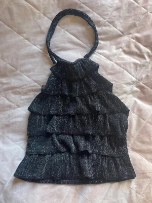 Glittrig svart halterneck topp H&M - Snygg svart halterneck topp från H&M Divided med glittrigt tyg och flera volanglager. Toppen är ärmlös och har en festlig känsla med sitt skimrande material och trendiga design. Perfekt för att sticka ut i mängden.
