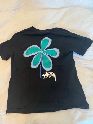 Svart Stüssy t-shirt med blå blomma - Svart t-shirt från Stüssy med stor blå och turkos blomma på ryggen och mindre blomma med logga på bröstet. Klassisk passform och rund hals. Perfekt för dig som gillar archive/streetwear