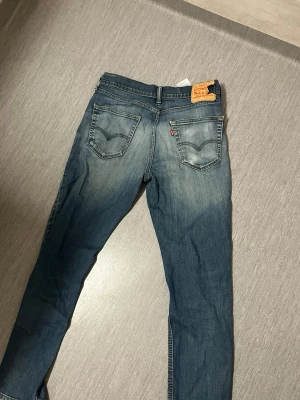 Levi's blå slim fit jeans - Säljer ett par klassiska blå Levi's jeans i slim fit-modell med snygga slitningar och femficksdesign. Jeansen har en ikonisk läderpatch bak i midjan och röda Levi's-tag på bakfickan. Perfekta för en avslappnad och trendig look.
