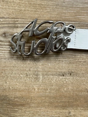 Vitt skinnbälte Acne Studios - Säljer ett vitt skinnbälte från Acne Studios med en unik silverfärgad buckle där loggan är formad i coolt script. Bältet har klassisk design med flera hål för justering och passar perfekt till jeans eller byxor. Strl 105 cm