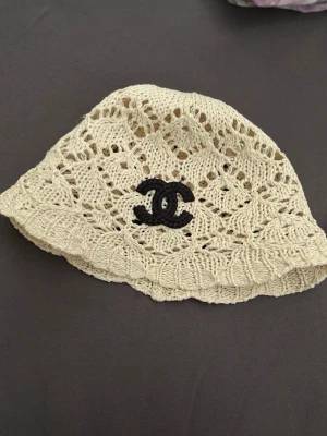 CH - Snygg beige bucket hat i virkat material från Ch  det klassiska svarta logotypbroderiet framtill. Hatten har ett luftigt och mönstrat virkat utförande som ger en trendig och somrig vibe. Perfekt accessoar för att lyfta din stil.