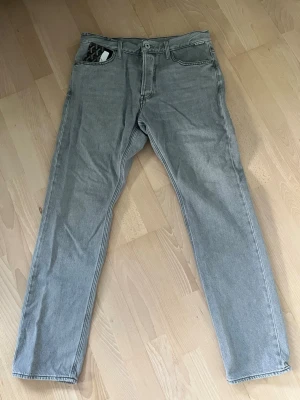 Grå jeans från G-Star  - Säljer ett par snygga grå jeans från G-Star  med klassisk femficksdesign och raka ben. Jeansen har en ljusgrå tvätt och detaljerad söm på bakfickan. Perfekta för en avslappnad och trendig stil.