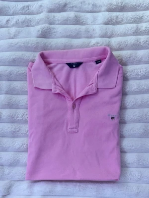 Rosa pikétröja från Gant - Snygg rosa pikétröja från Gant med klassisk krage och korta ärmar. Tröjan har två knappar vid halsen och en liten broderad Gant-logga på bröstet. Tillverkad i mjuk bomull som känns skön mot huden. En otrolig sommar piké 