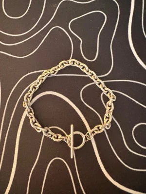 Silverfärgat kedjearmband med T-lås från skultuna - Stilrent armband i silverfärgad metall med grova kedjelänkar och ett trendigt T-lås från Skultuna. Perfekt accessoar för att lyfta din outfit med en modern touch. Passar både till lager-på-lager och som statement piece. PRISET ÄR INTE HUGGET I STEN!👍🏼