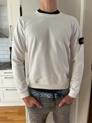 Vit sweatshirt från Stone Island - Vit långärmad sweatshirt från Stone Island med klassisk rund hals och ribbade muddar. Tröjan har den ikoniska logotypen på vänster ärm och är tillverkad i mjukt material som känns skönt mot huden. Perfekt för en clean och stilren look.