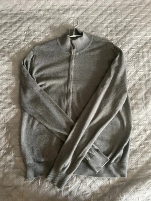 Grå ziptröja med hög krage - Snygg grå ziptröja med hög krage och ribbade muddar vid ärmslut och nederkant. Tröjan är tunnstickad och har en stilren look som passar perfekt till lager-på-lager. Dragkedja framtill för enkel på- och avtagning.
