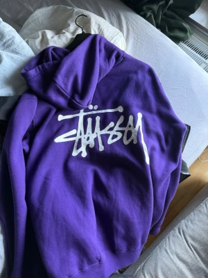 Lila Stüssy hoodie med tryck - Snygg lila hoodie från Stüssy med stort vitt logotryck på ryggen och mindre logga på bröstet. Klassisk design, Perfekt för dig som gillar streetwear och vill sticka ut med färg.