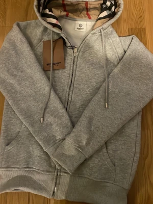 Grå zip hoodie från Burberry - Snygg grå zip hoodie från Burberry med klassiskt rutigt foder i huvan och dragsko. Tillverkad i mjukt bomullsmaterial och har fickor framtill. Perfekt för en avslappnad och trendig look.