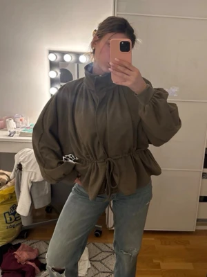 Olivgrön jacka från cider  - Säljer en olivgrön oversized jacka med hög krage och snörning i midjan. Jackan har långa ärmar och en avslappnad passform som ger en cool och trendig look. Perfekt att slänga på över en hoodie eller t-shirt.
