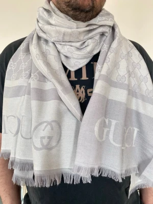 Ljusgrå halsduk från Guccii - Snygg ljusgrå halsduk från Guccii med klassiskt GG-mönster och stora logotyper. Halsduken har fransade kanter och är tillverkad i ett mjukt tyg som känns lyxigt. Perfekt accessoar för att lyfta din outfit.