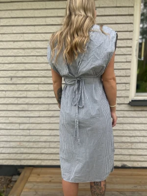 Randig knälång klänning med knyt från 2NDDAY - Snygg randig klänning i vitt och grått med knyt i midjan. Modellen är ärmlös och har en rak passform som ger en avslappnad look. Perfekt för varma dagar och enkel att styla med accessoarer. Använd max 2 gånger så nyskick! Storlek M