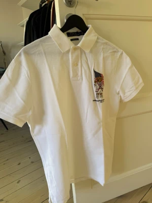 Vit pikétröja från Polo Ralph Lauren - Vit pikétröja med kort ärm från Polo Ralph Lauren. På bröstet finns en broderad nallebjörn som håller en amerikansk flagga. Klassisk krage med knappar och sportig känsla. Perfekt för en avslappnad och stilren look.