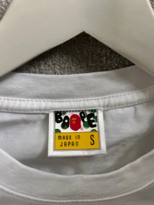 Vit Bape t-shirt med camomönster - Vit t-shirt från Bape med ikoniskt camomönstrat apansikte i rosa och rött på bröstet samt en liten Baby Milo-figur ovanpå. Klassisk passform och rund halsringning. Tillverkad i Japan, perfekt för dig som gillar streetwear.