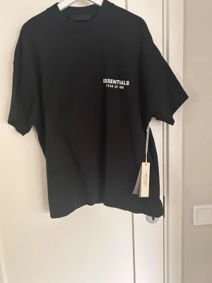 Svart Essentials Fear of God t-shirt - Svart t-shirt från Fear of God Essentials med vit logga tryckt på bröstet och ryggen. Klassisk rund hals och korta ärmar. Tillverkad i mjuk bomull för en skön och avslappnad passform. Perfekt för en stilren och trendig look. T-shirten sitter lite loose 