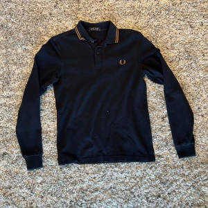 Svart långärmad piké från Fred Perry - Svart långärmad pikétröja från Fred Perry med klassisk krage och knappar framtill. Gula ränder på kragen och broderad logga på bröstet. Perfekt för en stilren och sportig look.