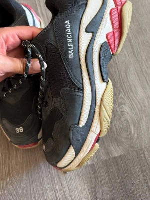Balenciaga Triple S svarta sneakers - Chunky svarta Balenciaga Triple S sneakers med flerlagrad sula i beige, svart och rött. Ovandel i mesh och mocka, vita loggan på sidan och storlek broderad framtill. Svarta och vita snören ger en sportig vibe. Perfekta för dig som gillar statement-skor.
