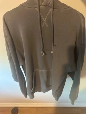 Grå hoodie med ficka och huva från Gymshark - En enkel och stilren grå hoodie med huva och dragsnören. Hoodien har en stor magficka framtill och synliga sömmar som ger en avslappnad look. Perfekt för dig som gillar en basic och bekväm stil. Materialet känns mjukt och skönt mot huden. Helt oanvänd oversized hoodie från Gymshark.