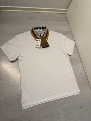Vit pikétröja med rutig krage Burberry - Vit pikétröja från Burberry med klassisk rutig krage i beige, svart, vitt och rött. Tröjan har korta ärmar och en stilren passform. Perfekt för dig som gillar exklusiva och tidlösa plagg med en twist. Fler bilder finns. Fraktar alltid samma dag eller dagen efter. Storlek M:)