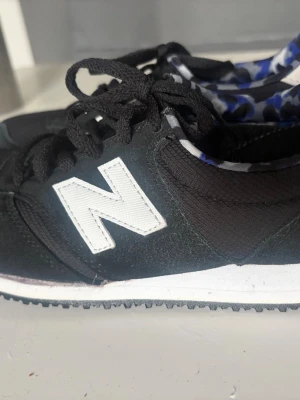 Svarta New Balance sneakers  - Svarta sneakers från New Balance med vit sula och stort vitt N på sidan. Sulan har lossnat i kanterna efter att jag tvättat den i tvättmaskinen, vilket jag inte borde ha gjort. Om du kan fästa den är allt bra.