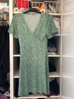 Grön blommig klänning Indiska S - Söt grön klänning från Indiska med vitt blommönster. Klänningen har v-ringning, korta ärmar och är i en kort modell. Perfekt för varma dagar och har en lätt och luftig känsla.