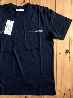 Svart Alexander McQueen t-shirt - Snygg svart t-shirt från Alexander McQueen med diskret logga broderad i rött och vitt på bröstet. Klassisk rund halsringning och korta ärmar. Tillverkad i mjuk bomull som känns skön mot huden. Perfekt för en stilren och trendig look.