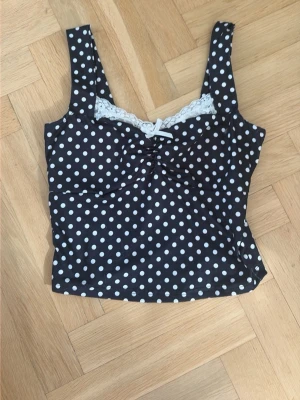 Svart prickig linnetopp med spets - Svart linnetopp med vita prickar och söt spetskant upptill. Toppen har breda axelband och en liten rosett framtill. Perfekt för en trendig och lekfull look.