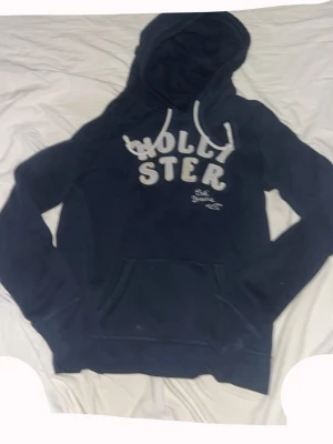 Mörkblå Hollister hoodie med tryck - Snygg mörkblå hoodie från Hollister med vit broderad logga och text på bröstet. Klassisk känguruficka framtill och vita dragsnören i huvan. Perfekt för en avslappnad och cool stil. Mjuk bomullsblandning som är skön att ha på sig.