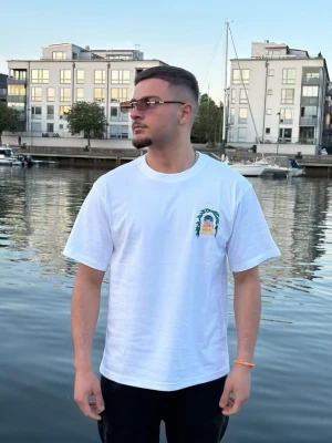 Vit t-shirt med tryck fram - Säljer en vit t-shirt med korta ärmar och ett färgglatt tryck på bröstet. T-shirten har en klassisk passform och rund halsringning. Perfekt till sommaren och enkel att matcha med olika outfits. 