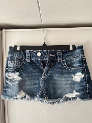 Blå jeanskjol med slitningar - Cool kort jeanskjol i blå denim med slitna detaljer och fransig nederkant. Kjolen har klassiska fickor fram och bak samt bälteshällor. Perfekt för en avslappnad och trendig look.