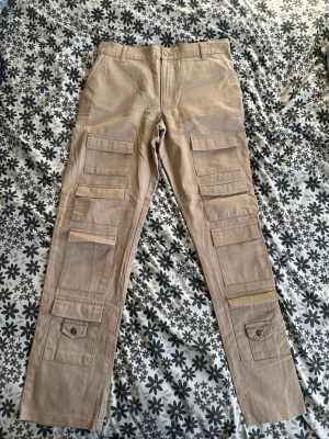 Beige cargobyxor med snygga fickor  - Säljer ett par beige cargobyxor med raka ben och flera stora fickor med lock och knapp framtill. Byxorna har bälteshällor och är tillverkade i ett slitstarkt bomullstyg. Perfekta för dig som gillar praktiska och snygga byxor med streetkänsla.