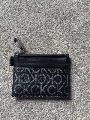 Svart korthållare/ plånbok från Calvin Klein - Snygg svart korthållare från Calvin Klein med CK-logga i mönster över hela framsidan. Plånboken har flera kortfack och en dragkedja med metall-detalj där det står Calvin Klein. Tillverkad i konstläder med stilren design. Pris kan diskuteras 