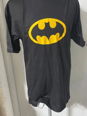 Svart Batman t-shirt med tryck - Cool svart t-shirt med det klassiska Batman-loggan i gult på bröstet. T-shirten har rund hals och korta ärmar, perfekt för dig som gillar superhjältar och vill sticka ut med en ikonisk look. Finns Stlk S, M , L och XL , 100% bomull, helt ny 