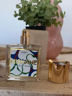 Nishane Tero Extrait de Parfum - Nishane Tero Extrait de Parfum i en stilren, dofter är använd och det finns 30 ml kvar av 50. Den är somrig jätte nishad och håller jätte länge. fyrkantig glasflaska med guldigt spruthuvud och lock. Flaskan har en färgglad etikett med gröna, blå och röda detaljer samt guldtext. Parfymen har en lyxig och modern design som sticker ut i parfymhyllan.
