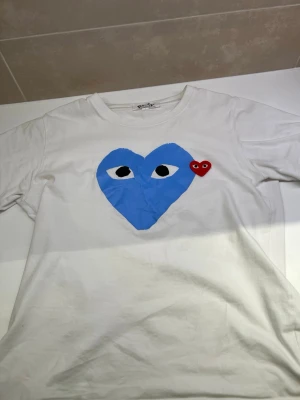 Vit t-shirt Comme des Garçons Play - Vit t-shirt från Comme des Garçons Play med stort blått hjärta med ögon och ett litet rött hjärta på bröstet. Klassisk rund hals och korta ärmar. Perfekt för dig som gillar streetstyle och coola prints.