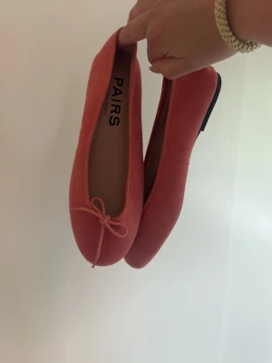 Korallrosa ballerinaskor från PAIRS - Snygga ballerinaskor i korallrosa från PAIRS. Skorna har en klassisk rund tå, liten rosett framtill och är gjorda i ett tygmaterial. 