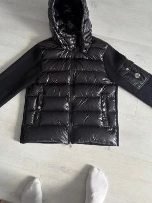 Moncler cardigan M - Snygg svart dunväst från Moncler med glansig finish, ribbstickade ärmar och huva. Västen har dragkedja framtill, två fickor med tryckknappar och en Moncler-logga på ärmen. Perfekt för kyliga dagar när du vill hålla stilen.