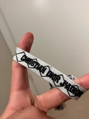 Vitt Christian Dior armband i tyg - Snyggt vitt armband från Christian Dior med svart broderad text och geometriskt mönster. Armbandet är gjort i tyg och har tofsar i ändarna för en cool och avslappnad look. Perfekt accessoar för att lyfta din outfit.