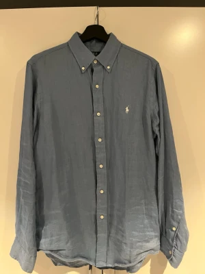 Blå linneskjorta från Polo Ralph Lauren - Snygg blå skjorta i linne från Polo Ralph Lauren med klassisk krage och knäppning framtill som är köpt på NK i Göteborg. Skjortan har lång ärm och den ikoniska broderade loggan på bröstet. Perfekt till sommaren.