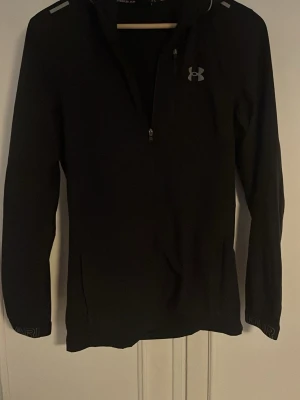 Svart träningsjacka från Under Armour - Svart långärmad träningsjacka från Under Armour med halv dragkedja framtill och reflekterande detaljer på axlarna. Jackan har en sportig look och är tillverkad i ett syntetmaterial som andas. Perfekt för aktiva dagar. Ändå dåliga är att dragkedjan är sönder så att jag har sytt där nere så man måste ta på den som en tröja 