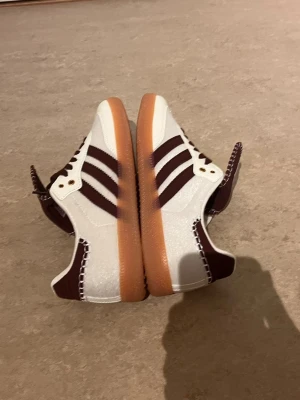 Adidas sneakers vita och vinröda - Snygga Adidas sneakers i vitt och vinrött med klassiska tre ränder på sidan. Skorna har en låg profil och en gummisula i ljusbrun färg. Ovandelen är i textil och syntet med detaljer i vinrött och vita sömmar. Perfekta för en stilren streetlook.