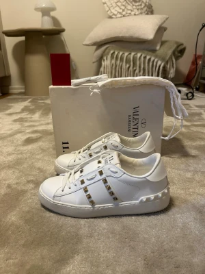 Vita Valentino Garavani sneakers - Snygga vita sneakers från Valentino Garavani med ikoniska nitar längs sidorna och på sulan. Skorna är i läder och har en klassisk låg modell med vita snören och diskreta detaljer som ger en lyxig vibe.