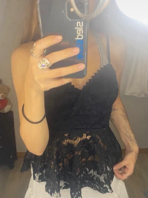 Svart spetsig topp - Snygg svart omlottopp med spetsdetaljer och volang nertill. Toppen har smala axelband och v-ringning, perfekt för att skapa en trendig look. Materialet är lätt och genombrutet med spets för en extra stylish känsla.