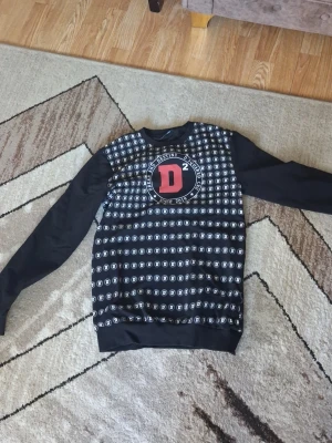 Svart Dsquared2 hoodie med D-mönster - Svart hoodie från Dsquared2 i storlek S med heltryckt D-mönster och stort rött D²-tryck på bröstet. Långa ärmar och ribbade muddar. Perfekt för dig som gillar streetwear och vill sticka ut med en unik design.