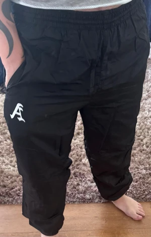 Svarta mjukisbyxor från Asap Rocky - Svarta mjukisbyxor med vit A-logga på vänster ben. Byxorna har resår i midjan och vid bensluten samt sidofickor. Tillverkade i ett glansigt, lätt material som ger en sportig vibe. Perfekta för chill eller streetwear.