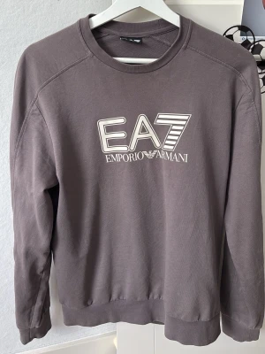 Emporio Armani Tröja - Snygg grå sweatshirt från Emporio Armani EA7 med tryckt logga framtill. Tröjan har rund halsringning, långa ärmar och ribbade muddar vid ärmslut och nederkant. Perfekt för en avslappnad och trendig stil. Använd 1 gång för test, bara höra av sig!