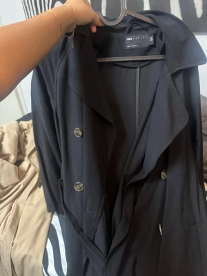 Svart trenchcoat från Odd Design - Stilren svart trenchcoat från Odd Design med klassisk dubbelknäppning och bred krage. Jackan har ett bälte i midjan och stora knappar framtill. Perfekt för dig som gillar en tidlös och trendig look.