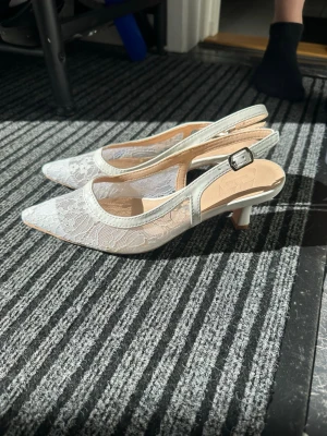 Vita spetsiga pumps med spetsdetaljer - Säljer ett par vita pumps med spetsig tå och låg klack. Skorna har fina spetsdetaljer och justerbar rem med spänne runt hälen. Perfekta för dig som gillar en elegant och romantisk stil. Materialet är en kombination av tyg och spets.
