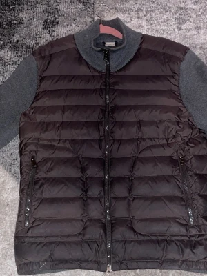 Svart dunväst från Moncler - Snygg svart dunväst från Moncler med quiltad design och gråmelerade stickade partier vid krage och axlar. Västen har dragkedja framtill och två fickor med dragkedja. Klassisk Moncler-logga på axeln. Perfekt för lager på lager och streetstyle.