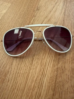 Vita aviator solglasögon med lila glas - Snygga aviator solglasögon med vit ram och lila tonade glas. Metallbågar med dubbla bryggor och klassisk pilotform. Perfekta för att lyfta din stil under soliga dagar.